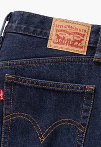 Un jean en denim bleu foncé présente un tab rouge distinctif avec le logo "Levi's", des coutures jaunes contrastantes et une pièce en cuir brun sur la taille.