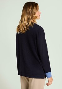 Cárdigan azul oscuro con un ajuste relajado. Presenta puños acanalados en azul claro y aberturas laterales. Fabricado con un material de punto suave.