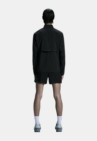 Sort atletisk jakke med en cropped ryg, parret med sorte shorts og grå løbesko. Simpelt design med minimale detaljer.