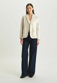 Blazer argenté brillant avec une coupe ajustée, revers crantés et deux poches avant, associé à un jean bleu foncé à jambes larges.