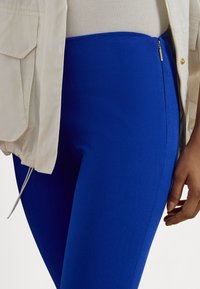 Modre elastične pajkice z gladko teksturo, s stranskim zadrgo in visokim pasom, kombinirane s svetlo bež jakno.