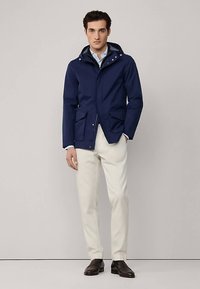 Mörkblå vattentät jacka med dragkedja och knappstängning, justerbar huva och flera framfickor, kombinerad med ljusa byxor och mörka skor.