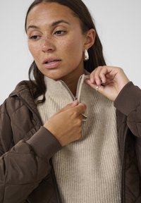 Brun quiltad jacka med huva över en beige ribbad polo. Modellen drar delvis upp dragkedjan på jackan. Silverörhängen och ring synliga.