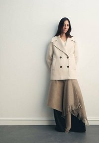 Cappotto in lana doppiopetto color crema con grandi bottoni neri, abbinato a una gonna a strati taupe e nera con bordi frangiati.