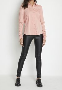 Femme portant une chemise rose à boutons avec des manches longues froncées, des leggings en cuir noirs et des mocassins noirs sur un fond blanc.