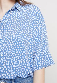 Kortärmad skjorta med knappar i ljusblått med ett oregelbundet prickigt mönster i vitt, med krage och avslappnad passform. Tillverkad av mjukt tyg.