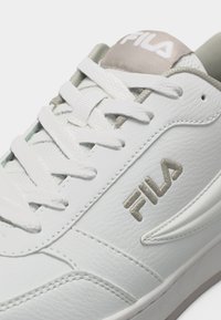 Baskets blanches FILA avec broderie de logo beige, cuir texturé et lacets blancs partiellement noués sur un fond clair.