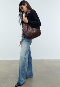 Femme en pull noir et jean large tenant un grand sac à bandoulière en cuir marron foncé, se tenant sur un sol en béton.