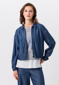 Denim jas met een ontspannen pasvorm, cropped ontwerp en een knoploos voorpand. Gedragen over een gestreepte shirt met een witte onderlaag.