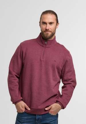 Bordeauxfarbener Pullover mit Viertelreißverschluss aus einem weichen, strukturierten Stoff. Mit hohem Kragen und schwarzem Logo auf der Brust. Getragen mit Jeans.