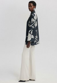 Femme portant une chemise florale noire et blanche surdimensionnée avec de larges manches, un pantalon blanc évasé, des escarpins noirs à bout pointu et de grandes boucles d'oreilles blanches.