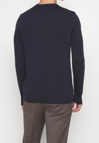 Man som bär en slim-fit mörk marinblå långärmad tröja och bruna byxor, sedd bakifrån mot en helt vit bakgrund.