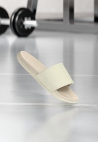 Ανοιχτό μπεζ σανδάλι slide με φαρδιά υφασμάτινη λουρίδα και επενδεδυμένο πάτο, κατασκευασμένο από μαλακό συνθετικό υλικό, τοποθετημένο σε φόντο γυμναστηρίου.