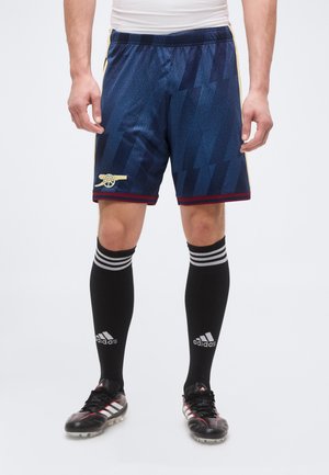 Uomo che indossa pantaloncini da calcio blu scuro con logo del cannone, calzini neri Adidas e scarpe da calcio nere, in piedi davanti a uno sfondo neutro.