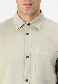 Cămașă verde deschis cu guler, având nasturi negri și un buzunar la piept. Fabricată dintr-un material texturat, cu un design simplu.