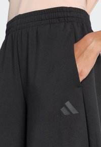 Pantaloni sportivi neri con cintura elastica, tasca laterale e un logo nero opaco sulla coscia sinistra. Tessuto morbido e design con vestibilità ampia.
