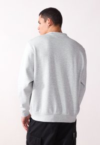 Felpa grigia con maniche lunghe, scollo rotondo e polsini a costine. Texture morbida, design casual, indossata sopra pantaloni cargo neri.
