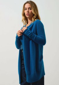 Cardigan in lana blu con polsini e orlo a costine, design aperto e texture morbida; abbinato a una maglia blu scuro e pantaloni di jeans.