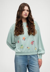 Mentazöld pulóver virágos hímezéssel, különböző színekben; laza szabás, bordás mandzsettákkal és derékpánttal. Kék farmerral párosítva.