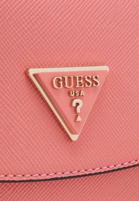 Borsa in materiale testurizzato rosa con un prominente emblema triangolare dorato recante la scritta "GUESS USA" e un punto interrogativo, contornato da cuciture nere.