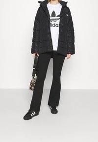 Svart puffjacka med huva, textil med struktur, dragkedjestängning; bärs över en vit Adidas-tshirt, kombinerad med svarta utsvängda byxor och svarta skor.