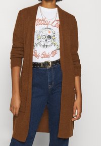 Brązowy długi kardigan o prążkowanej fakturze, z otwartym przodem i dwoma bocznymi kieszeniami, nałożony na białą t-shirtę z nadrukiem i ciemnoniebieskie dżinsy.