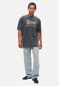 Grå bomulds T-shirt med et stort grafisk tekstdesign i ferskenfarve, parret med lyseblå slidte jeans med malingsplamager.