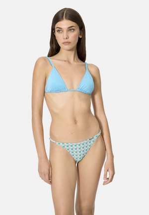 Mujer con cabello largo y castaño que lleva un top de bikini triangular azul claro y pantalones de bikini con estampado sobre un fondo liso y claro.
