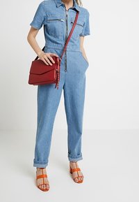 Žena v světlomodrém džínovém overal s krátkými rukávy a předním zipem, nese červenou křížovou tašku a má na sobě oranžové sandály na podpatku.