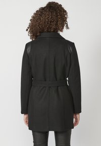 Cappotto nero con vita cinturata, texture liscia, spalle strutturate e dettagli a coste sulle spalle. Il design è aderente e moderno.