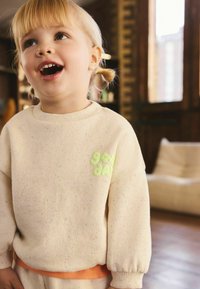 Suéter beige con motas de colores, con un texto verde que dice "buen día". Tejido suave, cuello redondo y puños fruncidos. Detalle de banda en la cintura naranja.