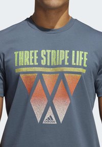 Šedé bavlněné tričko s grafickým vzorem ve formě oranžového a bílého trojúhelníku s textem "THREE STRIPE LIFE" a bílým logem adidas.