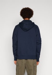 Felpa blu navy con cappuccio ampio, polsini a costine e vestibilità comoda, realizzata in cotone per il massimo comfort. Abbinata a pantaloni cargo verde oliva.