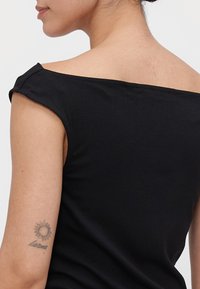 Sieviete, kura valkā melnu topu bez plecu siksnām, ar mazu saules tetovējumu un vārdu "karma" uz augšējā kreisā rokas.
