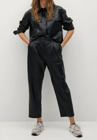 Conjunto de cuero sintético negro que incluye una camisa de botones con dos bolsillos en el pecho y pantalones de cintura alta, complementado con zapatillas grises.