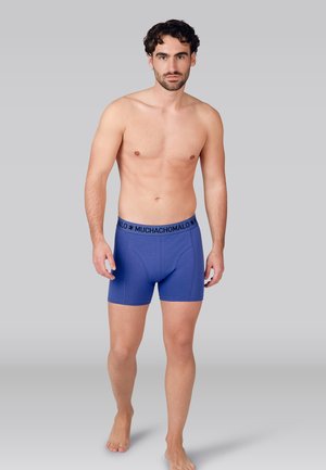 Jonge man met donker krullend haar, dragend blauwe boxershorts met "MUCHACHOMALO" tailleband, loopt blootsvoets tegen een effen grijze achtergrond.
