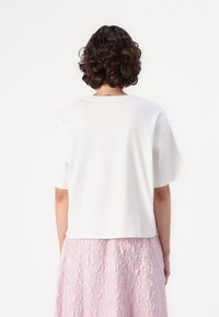 T-shirt en coton blanc à manches courtes et coupe décontractée, porté sur une jupe rose texturée. Le dos dévoile un design simple et minimaliste.