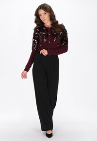 Burgunderfarbener Cardigan mit Paillettenverzierungen, Knopfverschluss, kombiniert mit hochgeschnittenen schwarzen Hosen und schwarzen High Heels, auf einem weißen Hintergrund.