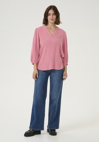 Blouse rose texturée avec un col en V et des manches trois-quarts, associée à un jean bleu à jambes larges et des chaussures à plateforme noires.