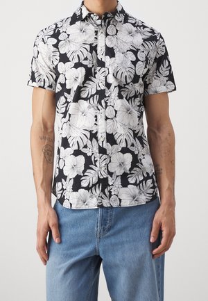 Mann trägt ein schwarzes kurzärmeliges Hemd mit weißem tropischem Blumen- und Blättermuster, kombiniert mit hellblauen Jeans.