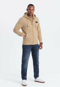 Sudadera con cremallera beige, con cordones, bolsillo en el pecho y puños acanalados, combinada con jeans azul oscuro y zapatillas blancas sobre un fondo liso.