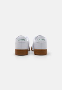 Bijele kožne tenisice s smešnim gumenim đonom, zaobljenog oblika i zelenim "LACOSTE" brendingom na poleđini. Minimalistički dizajn bez uzoraka.