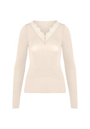 Jonge vrouw in een crèmekleurige top met lange mouwen en kanten V-hals, houdt haar handen samen en kijkt iets naar rechts tegen een effen achtergrond.