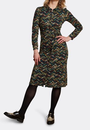 Kobieta w midi sukience na długi rękaw z wzorem i paskiem, czarnych rajstopach oraz czarno-beżowych sznurowanych butach, pozująca z jedną wyciągniętą nogą.