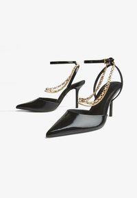 Črne lakirane visoke pete s ostro konico, z ankelnimi trakovi in zlatimi verižnimi detajli na srednjem delu stopala. Tanek stiletto peti.