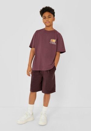 Jongen met krullend haar, draagt een paars T-shirt met een oranje afbeelding, bijpassende korte broek, witte sokken en witte sneakers, staat met één hand in zijn zak.