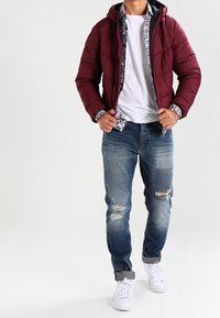 Maroon puffjacka med huva, bärs över en vit T-shirt. Slitna blå jeans med en blekt tvätt och vita sneakers.
