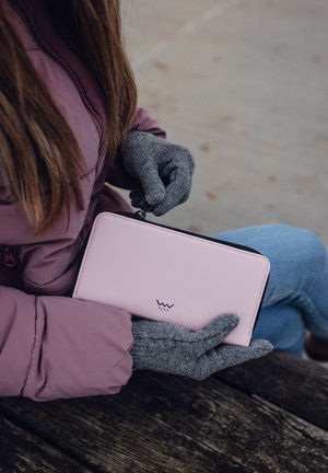Pink leather wallet with a zip closure, featuring a logo. Gray gloves hold the wallet while sitting on a wooden surface.
