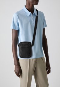 Geantă de umăr neagră cu o textură netedă, av�ând un buzunar frontal cu fermoar și o curea cu logo, purtată peste un tricou polo bleu deschis și pantaloni bej.