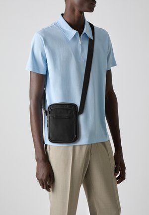 Black crossbody bag with a smooth texture, featuring a front zip pocket and branded strap, worn over a light blue polo shirt and beige pants.